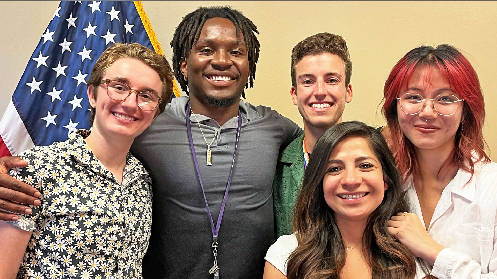 5 diverse students from a prior cohort pose in front of the American flag