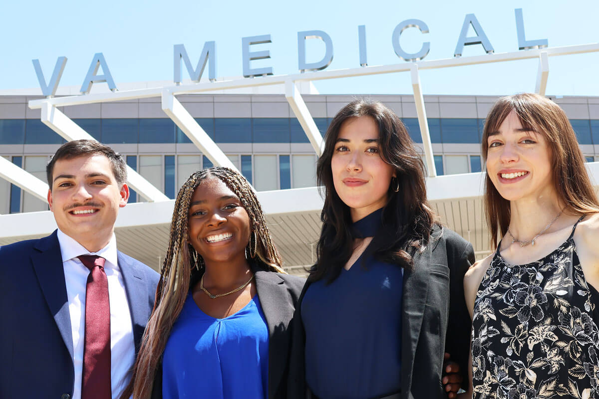 4 diverse students from a prior cohort pose in front of the VA Medical Center