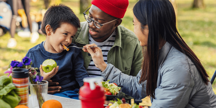 Father holds child on lap as mother feeds the child nutritious food