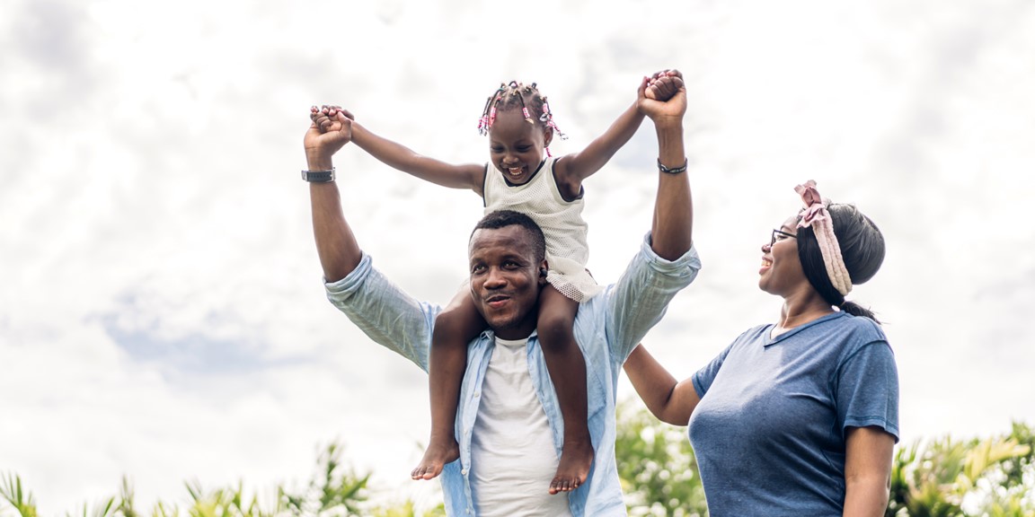 A father holds his daughter on his shoulders, lifting up their arms, while his wife looks on lovingly